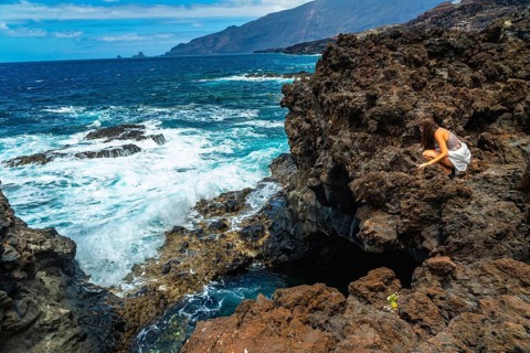 Woman crouching on dark volcanic rocks, observing the vibrant turquoise ocean water crashing