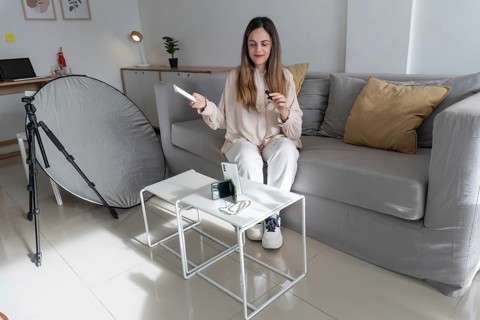 Woman sitting on couch holding smartphone and small microphone, camera tripod and silver reflector