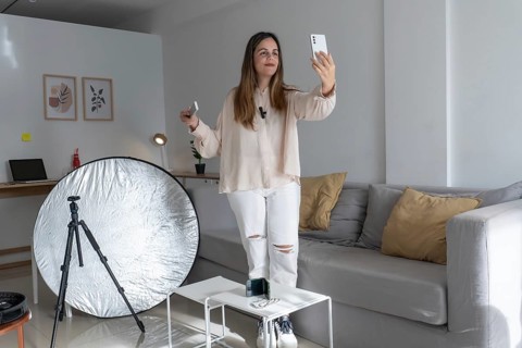 Full length shot of woman standing and taking a selfie with smartphone, tripod and reflector set up