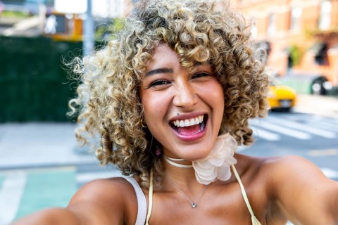 Young woman with curly blonde hair smiling broadly and taking a selfie, enjoying an urban lifestyle