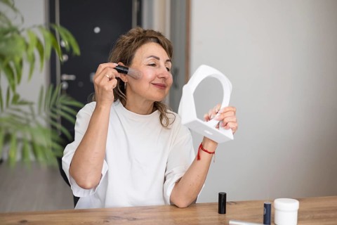 A mature pretty woman with medium-length hair is applying makeup using a brush while gazing into a