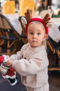 A young Caucasian girl with blonde hair wears a reindeer headband. She holds a small Santa toy in a