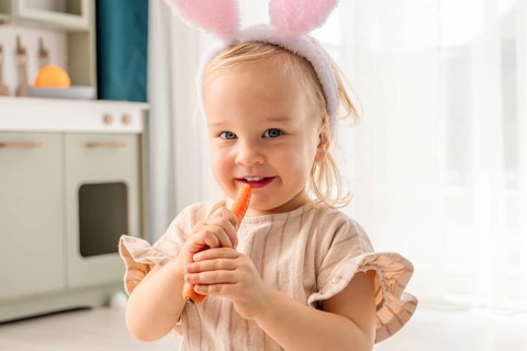 A young girl in bunny ears holds a carrot and smiles. She enjoys the Easter holiday while standing