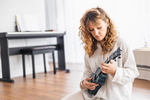 Person sits with a ukulele near a piano in a room with wooden floors