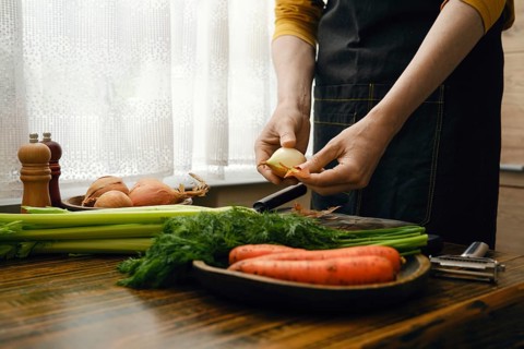 A person peels an onion while preparing vegetables in a kitchen. Carrots, celery, and herbs are
