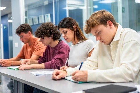 Group of young students sitting at desks in a modern classroom, intensely focusing and writing on