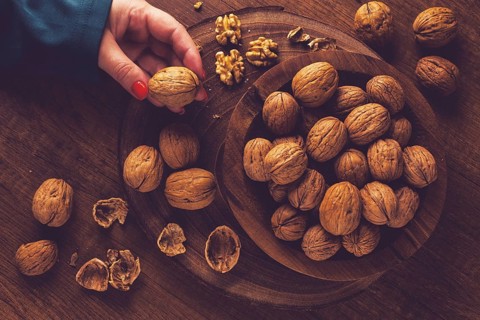 A woman's hand taking a walnut from a wooden bowl filled with walnuts. There are shells and walnuts