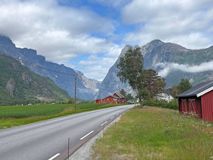 A road leads through a green valley flanked by mountains and red houses under a partly cloudy sky,