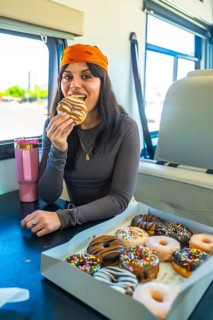 Young woman smiling and taking a bite from a glazed donut, sitting at a table with an open box of