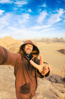 Smiling male tourist traveler wearing hooded traditional brown farwa cloak taking selfie making