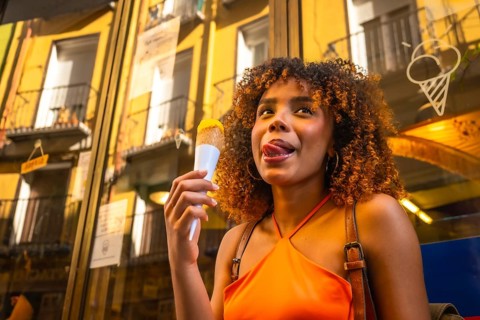 Young woman with curly afro hair savoring an ice cream cone on a sunny city street, smiling and
