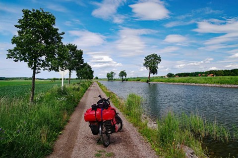 A loaded bicycle stands on a narrow path along a river under slightly cloudy sky, bicycle tours,