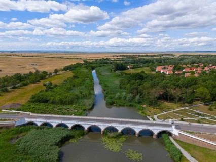 Wide landscape with a river and a bridge surrounded by fields and a village under a cloudy sky,