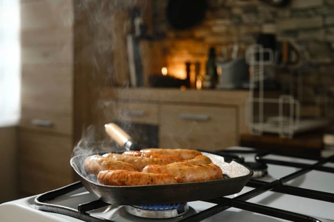 Banger sausages frying in cast-iron griddle pan on stovetop in kitchen. Smoke from melted fat rises