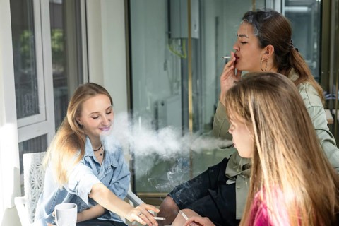 Three friends are sitting at a table in a cafe during the afternoon. They are smoking cigarettes