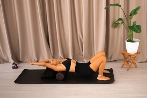 Woman stretching her back using a foam roller on a yoga mat next to indoor plants