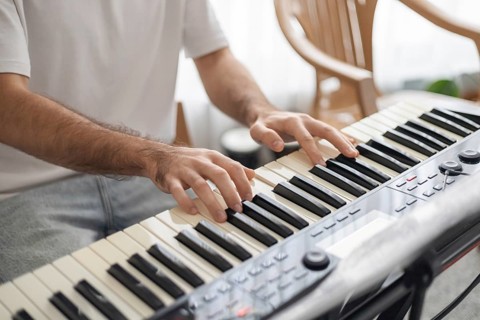 A young man with dark hair plays a keyboard at home. The setting is cozy, with wooden chairs and
