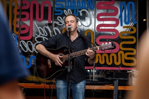 A middle-aged Caucasian man with short hair sings into a microphone while playing an acoustic