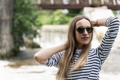 A woman in sunglasses and a striped top holds her hair near the river with her hands, A woman