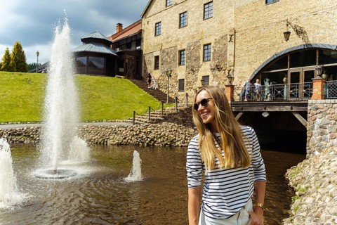 A long-haired woman in a striped jumper and sunglasses laughs next to a fountain in the summer, A