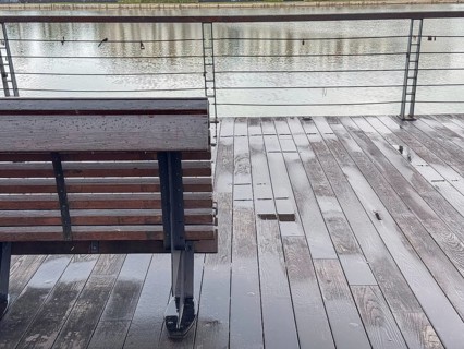 A wooden bench on a wet deck overlooking a calm body of water. The scene is tranquil and reflects a
