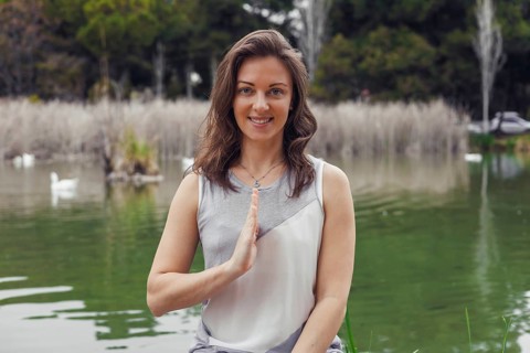 A young Caucasian woman with brown hair sits by a serene lake, practicing yoga. She smiles and