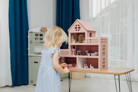 Child in a blue dress playing with a dollhouse set on a wooden table in a bright room