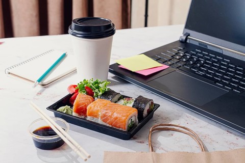 A person enjoys lunch at a workspace with sushi, salad, and coffee. A laptop is open with sticky