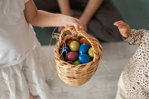 Two young girls holding a woven basket filled with colorful eggs. The scene captures a joyful