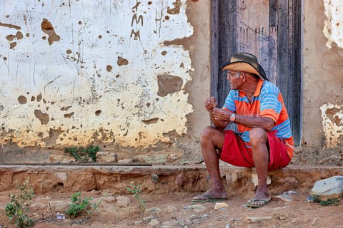 Elderly man sits thoughtfully in front of a faded wall on a village path, a man sits on a wall in