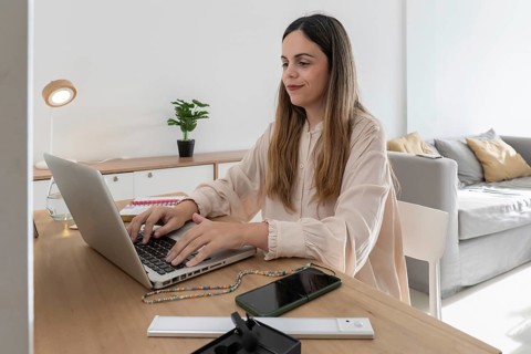 A content creator working at a wooden desk, typing on a laptop with a smartphone and headphones on
