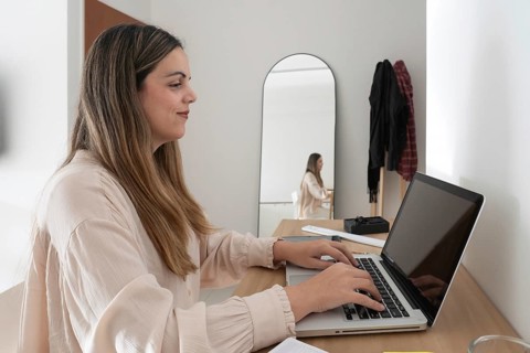 Close side profile of woman working on laptop at desk, reflection visible in tall arched mirror,