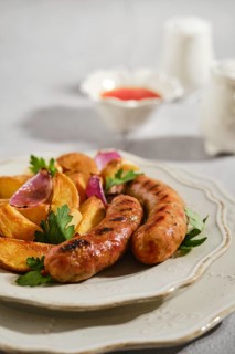 Sausages cooked to golden brown sit next to crispy potato wedges on white plate. Fresh herbs add