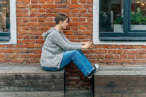 Woman 40s in knit cardigan using smartphone on wooden bench at brick building, A woman in casual