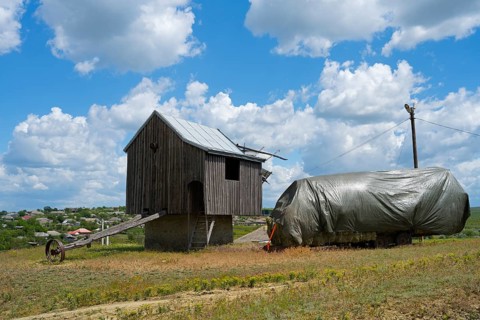 Wooden windmill next to a structure covered with tarpaulin in a field under a blue sky, surrounded