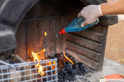 A gloved hand pours blue liquid onto burning charcoal in a stone barbecue grill. Flames rise from