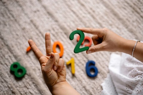 Hands displaying a peace sign and holding a green number two over a rug with numbers