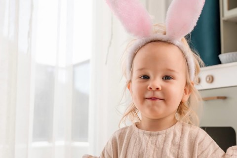 A toddler girl wearing bunny ears is celebrating Easter. She shows a joyful expression while