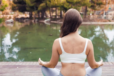 A young Caucasian woman with long brown hair sits cross-legged by a serene lake, practicing