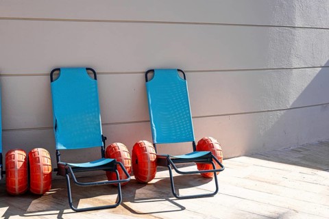 Four blue beach loungers with orange inflatable rings are lined up against a beige wall. The scene