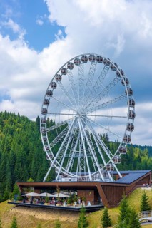 A large Ferris wheel set against a forested hillside under a partly cloudy sky
