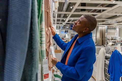 A young Black man examines fabric samples in a home goods store. He wears a blue coat and looks