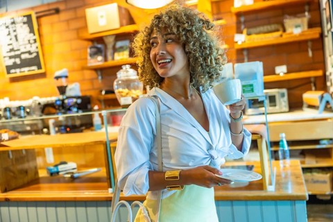 Smiling woman with curly hair standing in a modern coffee shop, holding a cup and saucer,