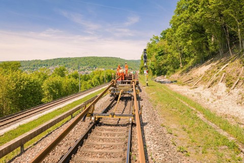 Workers in orange work on railroad tracks surrounded by trees on a sunny day, Leonhard Weiss