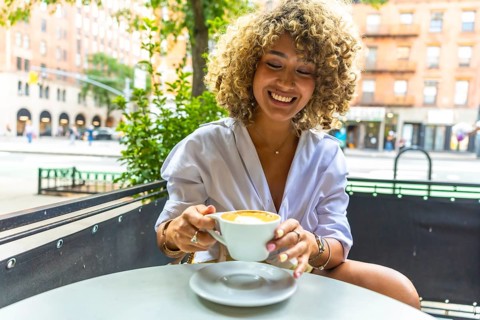 Happy young woman with curly hair sitting at an outdoor cafe table, smiling and holding a coffee