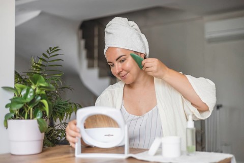 A young Caucasian woman with brown hair in a towel smiles while applying gua sha on her face. A