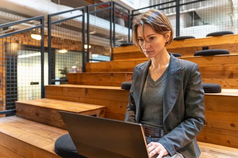 Professional woman in blazer working on laptop on wooden steps in modern coworking space