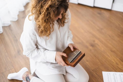 A woman engages with a kalimba, seated on a wooden floor with music sheets nearby
