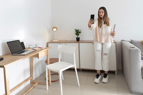 Full length shot of woman holding phone up while filming herself, standing in tiled living room