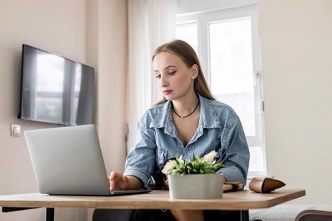 Young Caucasian woman with long brown hair working on a laptop at a wooden table. A potted plant is